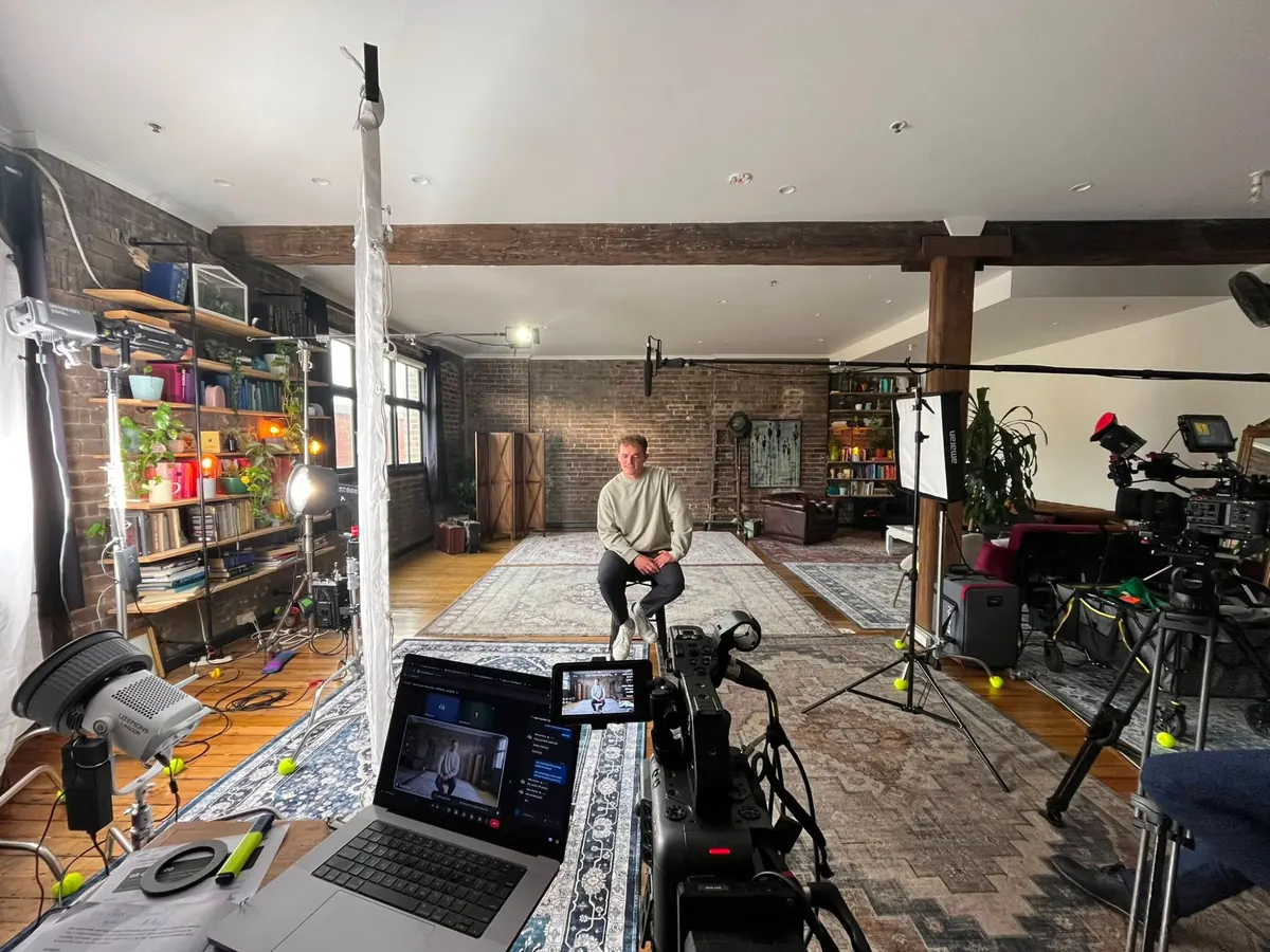 An interview set in a brick-walled loft with cinema cameras, lights, a boom microphone and a laptop running the remote video link