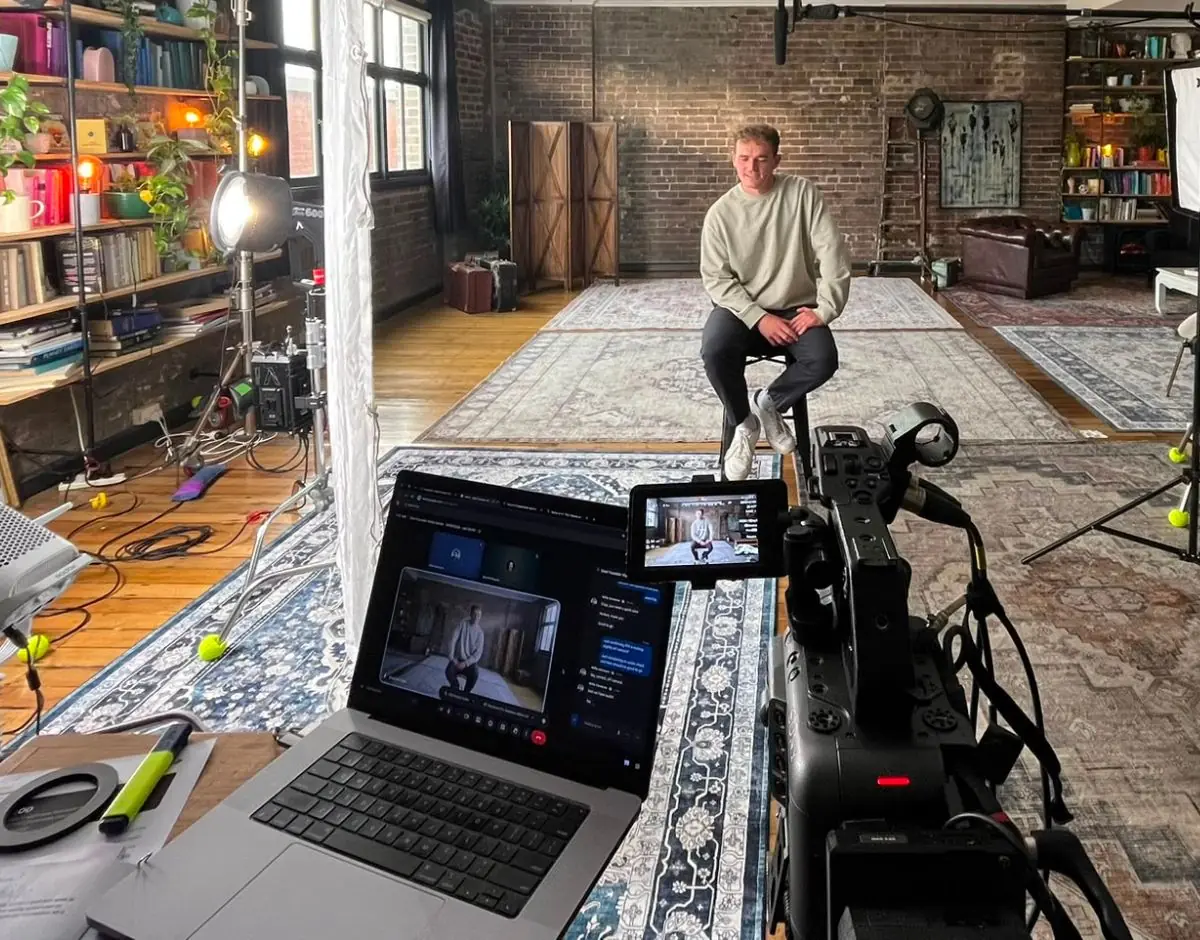 A laptop running a remote video call beside a cinema camera and its monitor, with the interviewee seated in a brick-walled loft behind