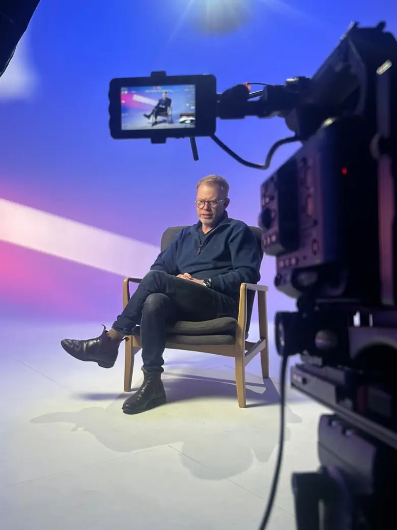 A studio camera and its on-board monitor framing a seated presenter against a blue-lit backdrop