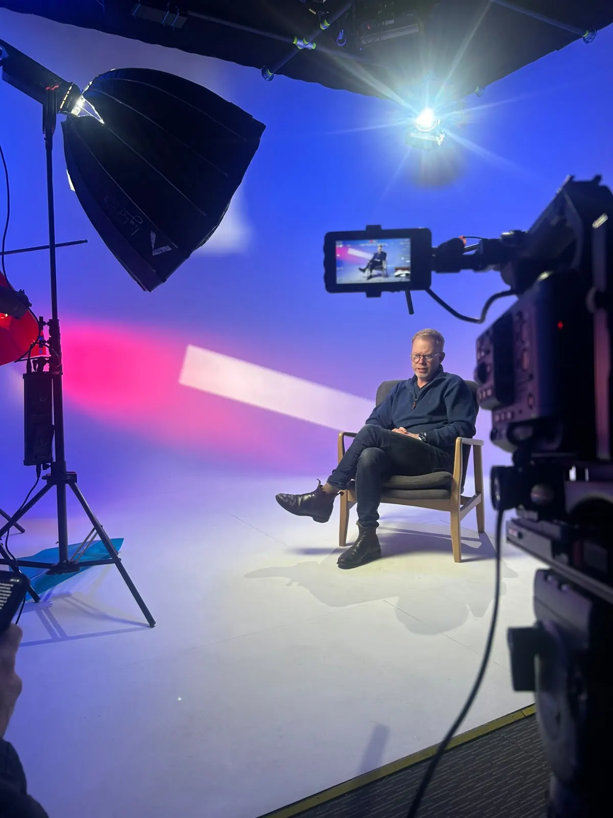 A presenter seated in a studio lit in blue and magenta, framed by a softbox light and a camera with an on-board monitor
